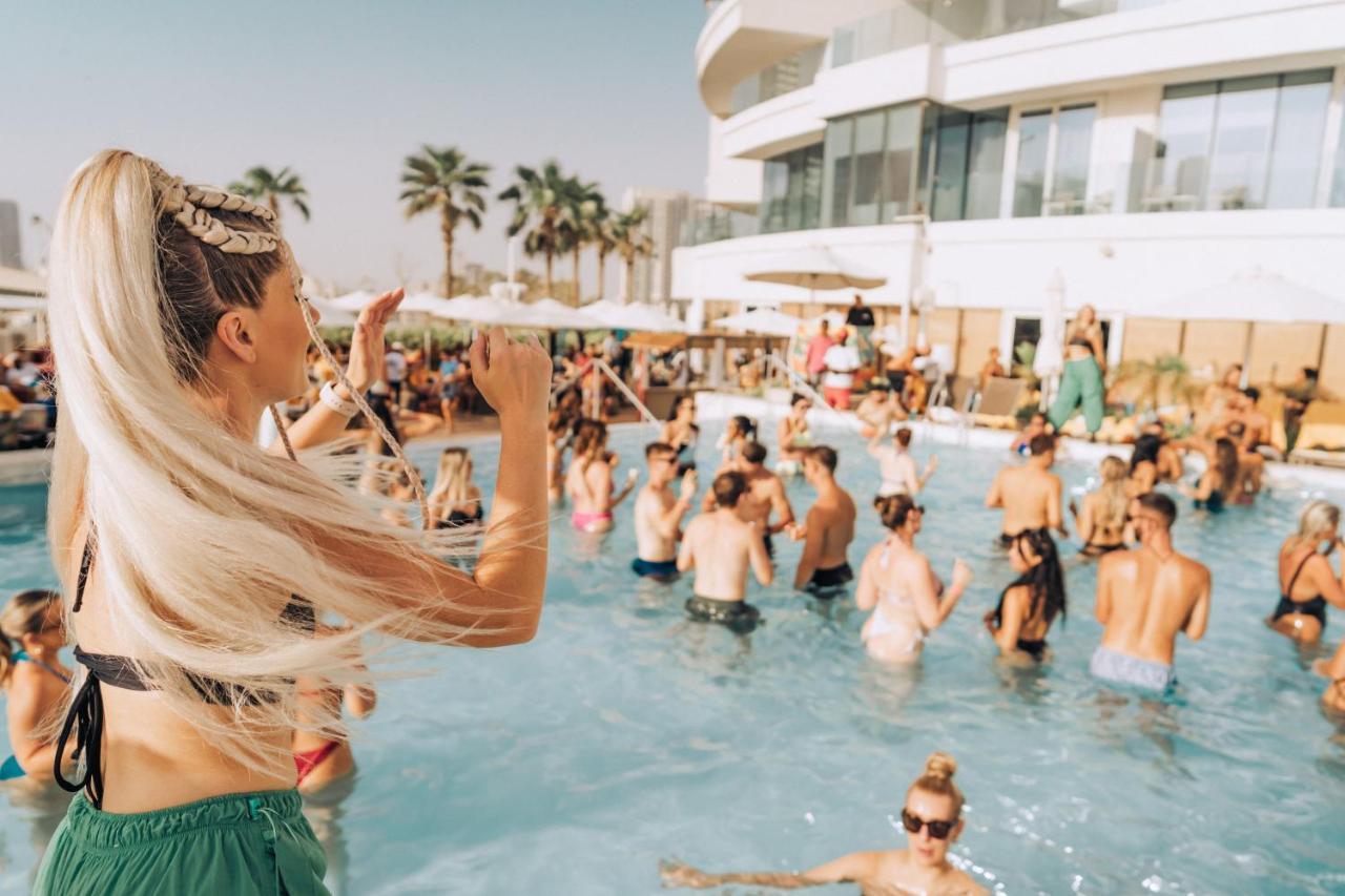 Five Jumeirah Village 두바이 외부 사진 The photo features a lively pool party scene. In the foreground, a woman with long, blonde hair is seen raising her arm as if enjoying the moment. The pool is filled with many people, some swimming and others socializing. The atmosphere appears festi