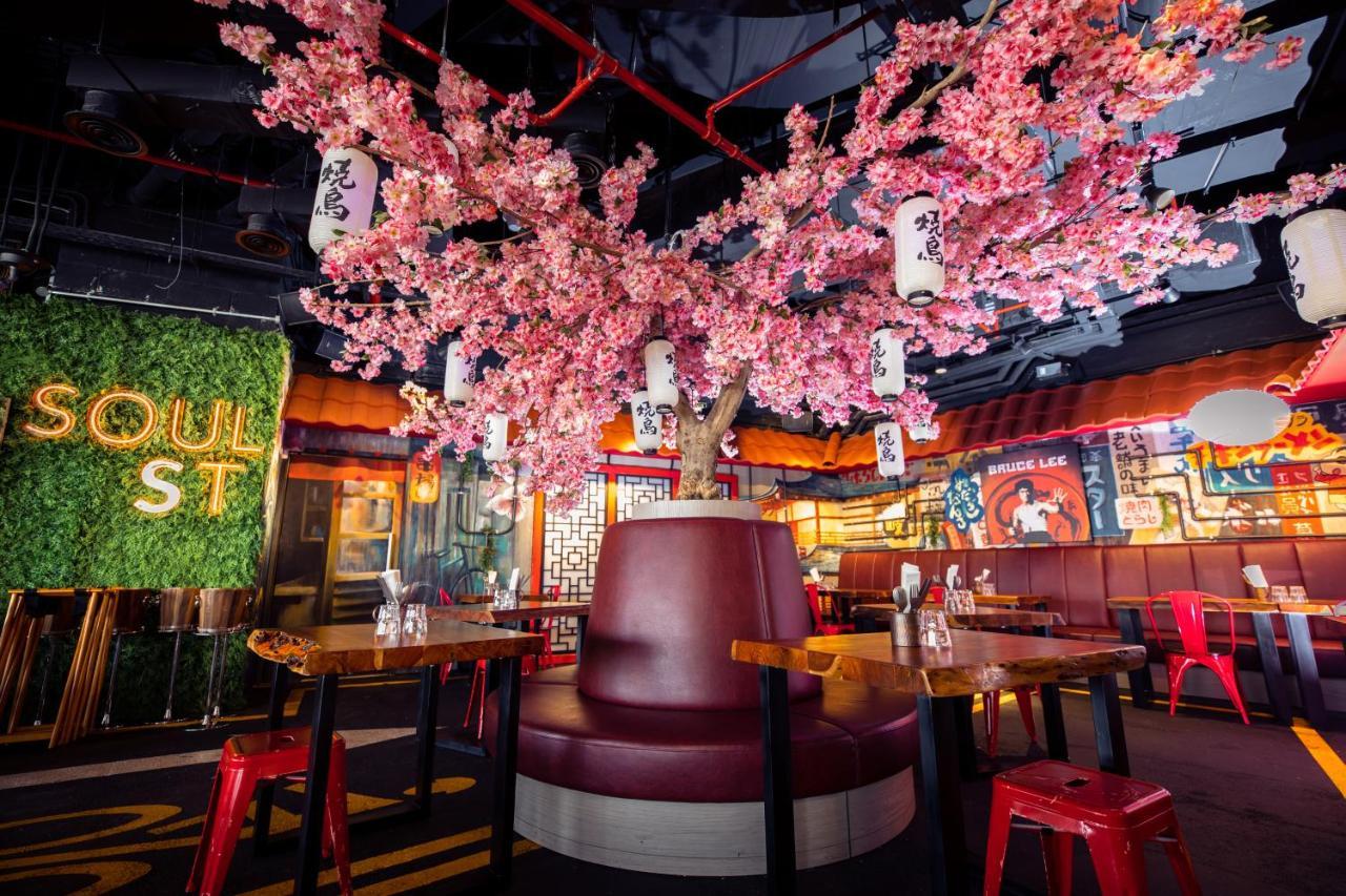 Five Jumeirah Village 두바이 외부 사진 The photo shows a vibrant and modern restaurant interior. At the center, there is a large cherry blossom tree adorned with pink flowers and hanging lanterns, creating a lively atmosphere. Surrounding the tree is a circular booth with maroon upholster