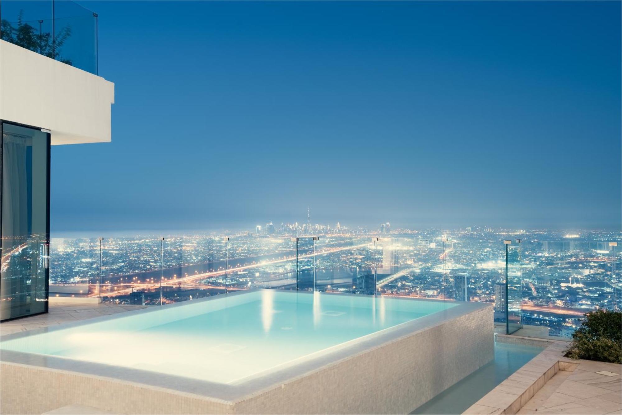 Five Jumeirah Village 두바이 외부 사진 The photo showcases a luxurious infinity pool situated on an elevated terrace, overlooking a sprawling cityscape at night. The pool's water has a serene glow, reflecting the city lights below, which shimmer and create a vibrant backdrop. The architec