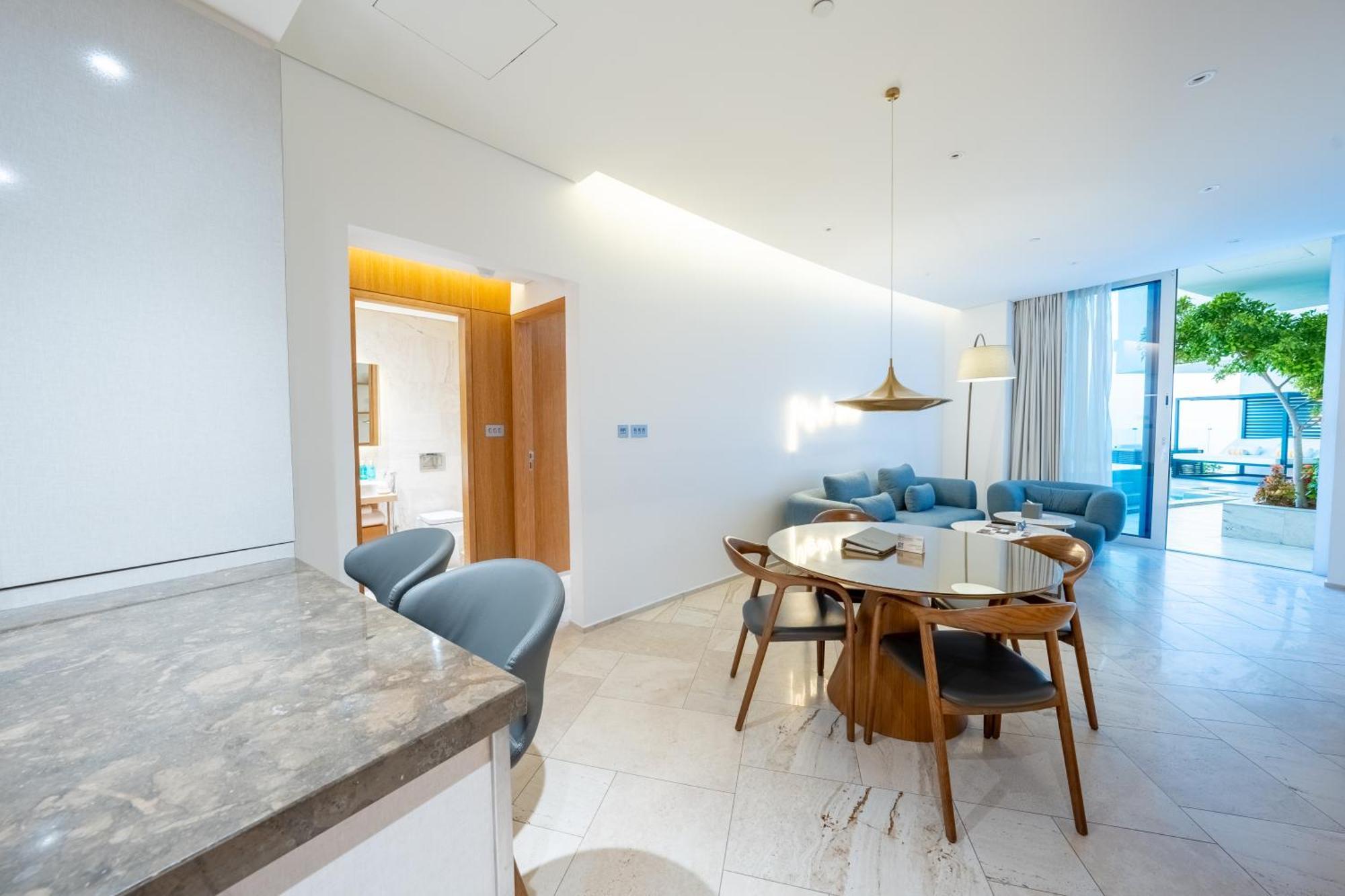 Five Jumeirah Village 두바이 외부 사진 The photo shows a modern living space that combines a dining area and a lounging area. On the left, there is a kitchen counter with a marble surface and several gray chairs around it. In the center, there is a round dining table made of wood with sev