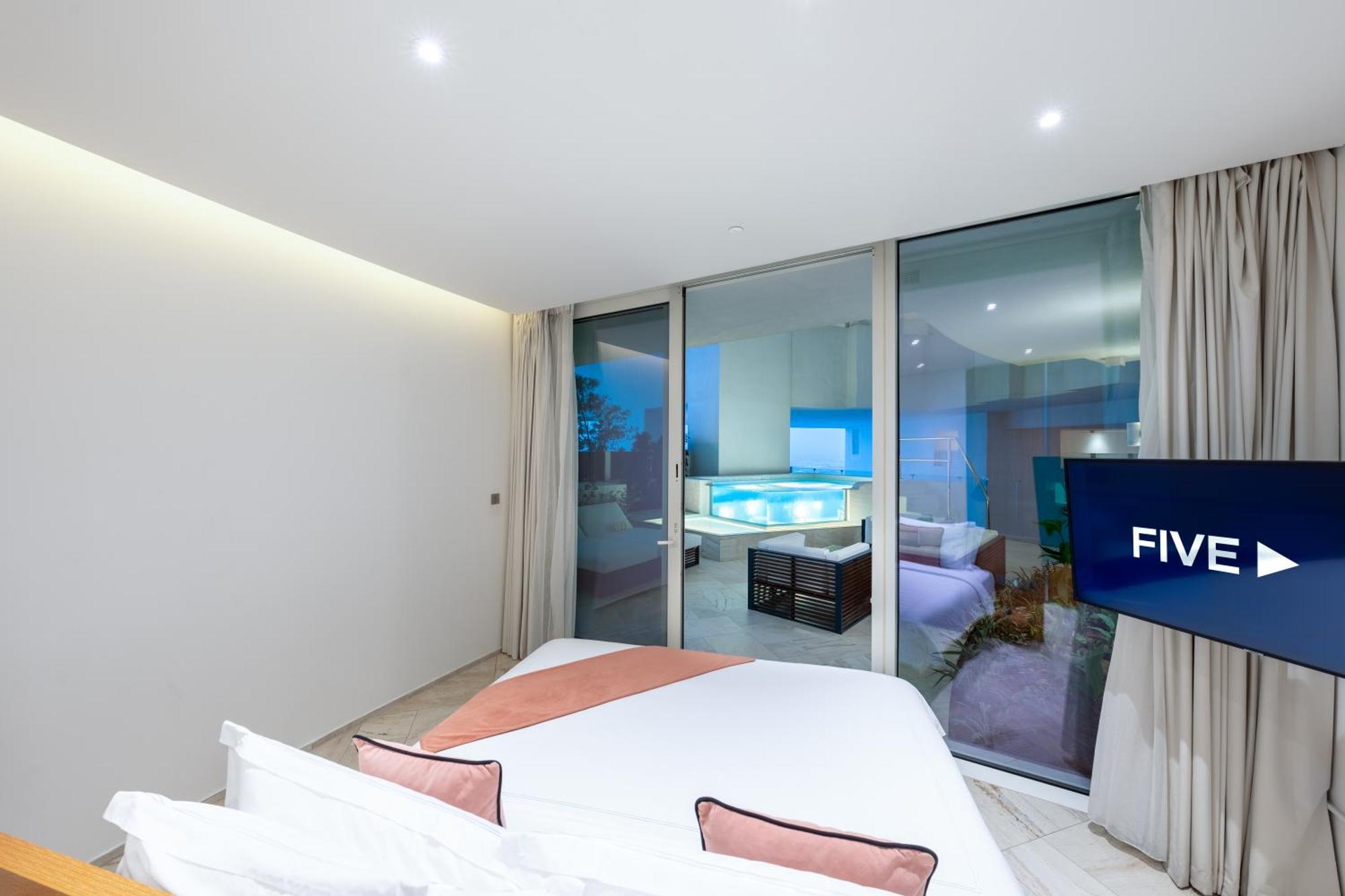 Five Jumeirah Village 두바이 외부 사진 The photo shows a modern, stylish bedroom featuring a large bed with light-colored bedding and decorative pillows. There are large glass windows that open up to a view of an outdoor area, likely overlooking a pool or patio. The room has neutral-color