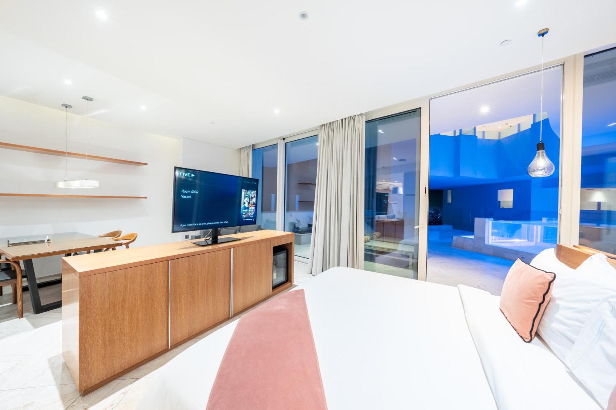 Five Jumeirah Village 두바이 외부 사진 The photo depicts a modern hotel room with a clean and minimalist design. A large bed is shown, topped with white linens and decorative pillows. Opposite the bed, there is a wooden TV stand with a flat-screen television. The room features large windo