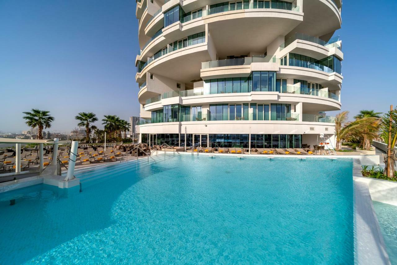 Five Jumeirah Village 두바이 외부 사진 The photo depicts a modern building with a distinctive architectural design, featuring multiple curved balconies. In front of the building, there is a large, inviting swimming pool with clear, turquoise water. Surrounding the pool are loungers for re