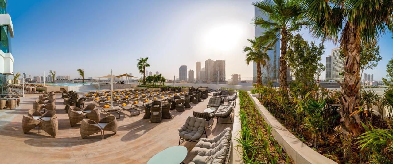 Five Jumeirah Village 두바이 외부 사진 The photo showcases a luxurious outdoor seating area, likely part of a resort or hotel. In the foreground, there are well-arranged lounge chairs and seating options, some with cushions, placed on a tiled surface. The area is bordered by lush green pl